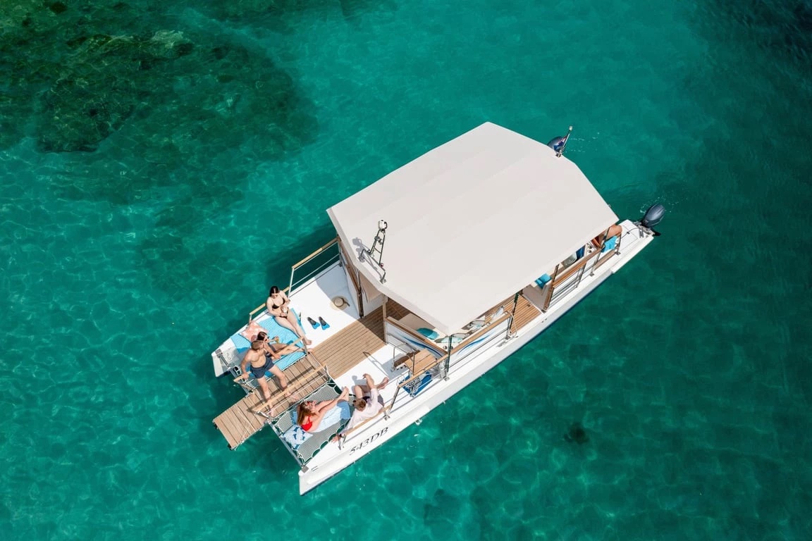 Aerial view of Catamaran X at anchor in turquoise shallows, guests relaxing on the bow deck beside the shaded canopy