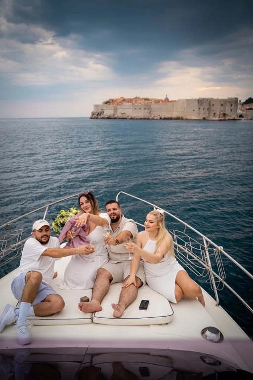 Four guests raising glasses on the Galeon 330 Fly bow sunpads, the old town walls across the water