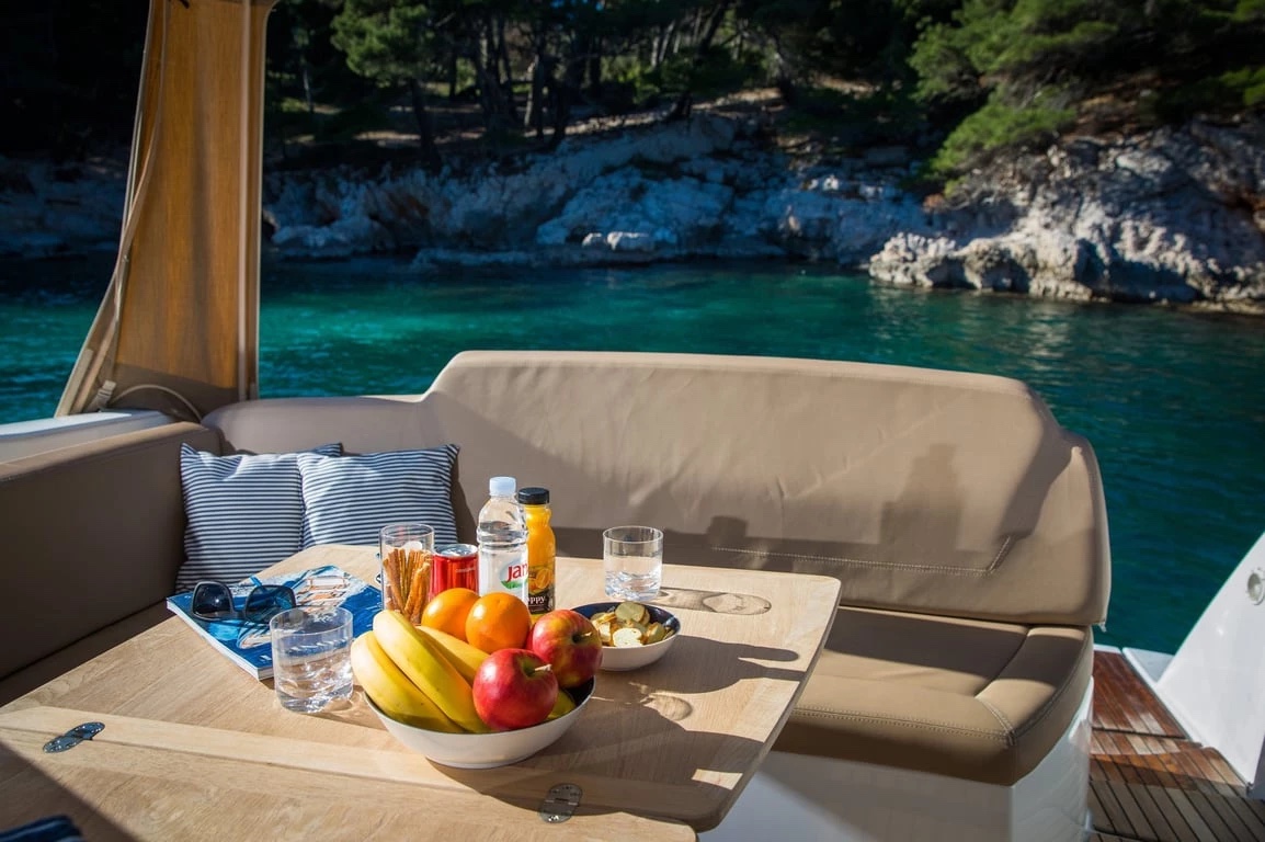 Close-up of the Jeanneau Leader 10's aft table — fruit bowl, drinks and sunglasses with a rocky cove beyond the stern