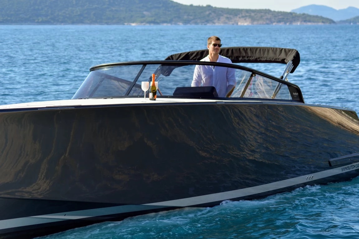 Skipper in a white shirt at the helm of the VanDutch 32, champagne on the foredeck, glossy dark bow filling the frame